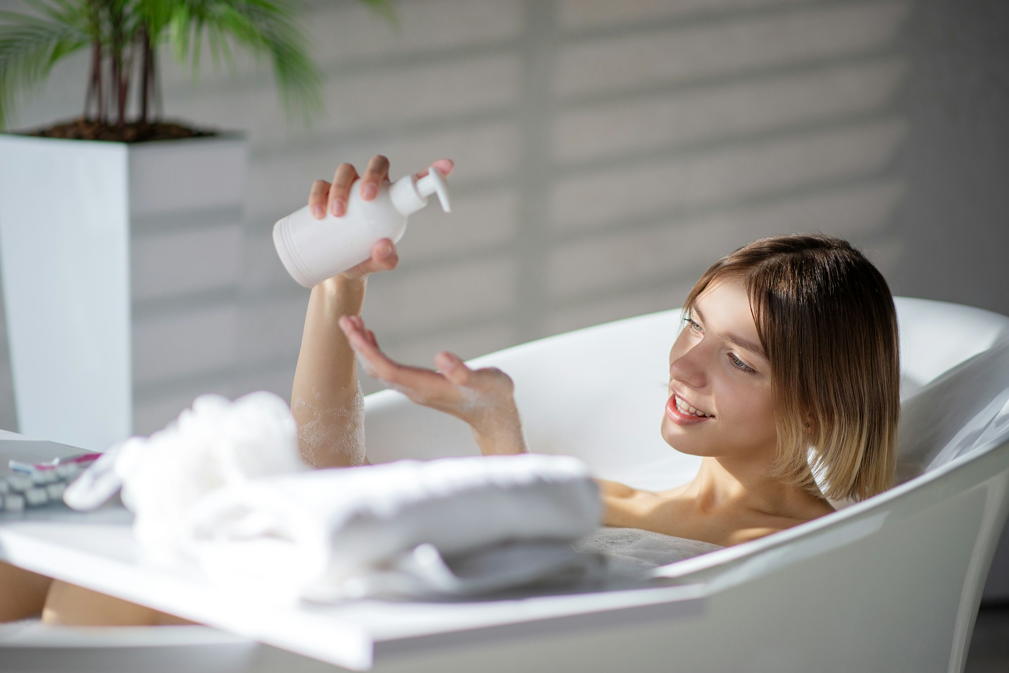 Beautiful woman pouring body wash on sponge, while resting in bathtub.